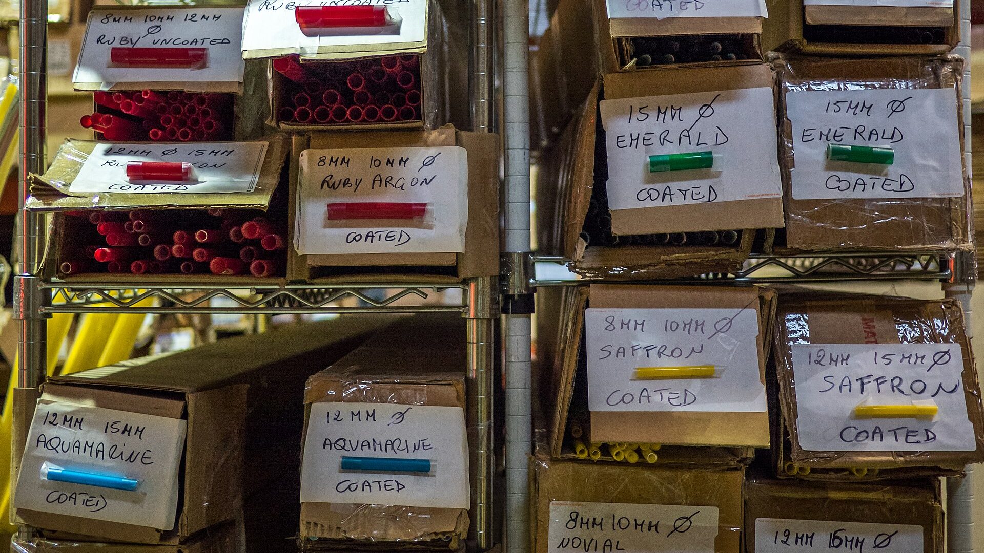 Warehouse shelving with boxes of uncoated and coated neon glass tube stock, labeled by color and diameter